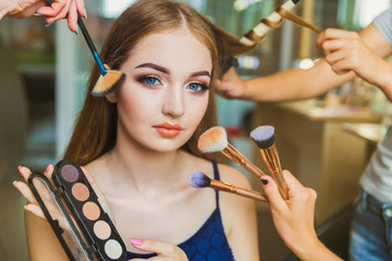 Portrait of the happy young woman who is sitting indoors and makeup artist doing makeup her and hairdresser doing the hairdo her