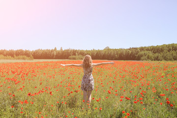 Beautiful girl is standing on summer field full of red poppy flowers in the grass. Sunny day with green lawn background. Happy woman in rustic dress holds hands sideward. The wind in her hair.