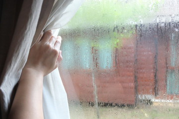 Woman's hand opening curtains in the bedroom with rain droplet on the glass, natural light and blurred garden background with text space.