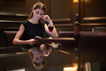 Young elegant woman with smartphone texting and having wine while sitting by table in restaurant