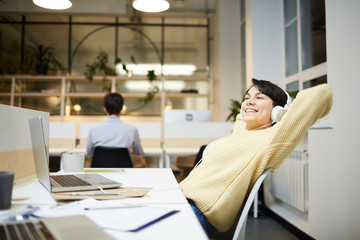 Young office manager with headphones enjoying her favorite music by workplace in front of laptop