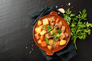 Meat stewed with potatoes, carrots and spices in ceramic pot on black background .