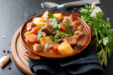 Meat stewed with potatoes, carrots and spices in ceramic pot on black background .