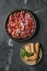 Antipasto meat platter assorti of sliced jamon, salami, chorizo sausage in terracotta tray with bread and greens over black marble background. Flat lay, space