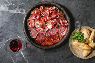 Antipasto meat platter assorti of sliced jamon, salami, chorizo sausage in terracotta tray with bread and glass of red wine over black marble background. Flat lay, space