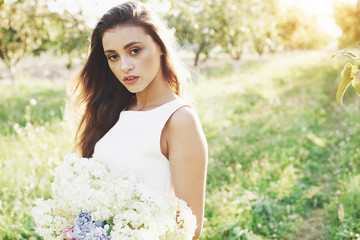 A beautiful young girl in a white light dress and a bouquet of summer flowers lays a fine day in the garden