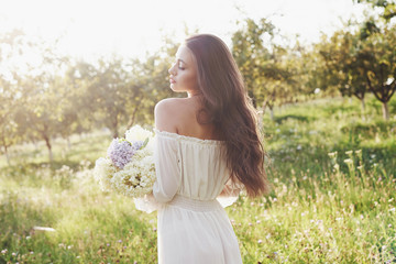 Fototapeta na wymiar A beautiful young girl in a white light dress and a bouquet of summer flowers lays a fine day in the garden
