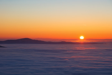 Background photo of low clouds in a mountain valley, red orange sky. Sunrise or sunset view of mountains and peaks peaking through clouds. Winter alpine like landscape of high siberia 