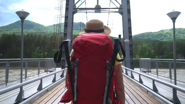 The woman the traveler with a red backpack goes along the cable-stayed bridge through the mountain river.