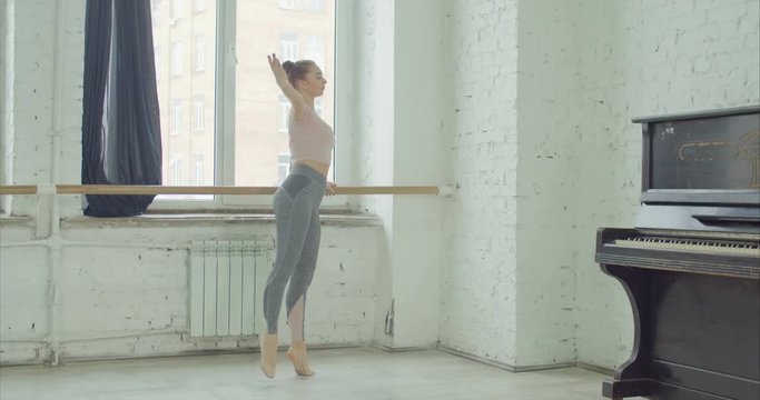 Elegant ballet dancer in sport cloches practicing developpe choreographic exercises at barre in rehearsal room. Graceful ballerrina performing classic dance moves during training in ballet studio.