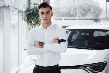 Fototapeta premium A handsome man is a buyer standing next to a new car at the dealer center and looking at the camera.