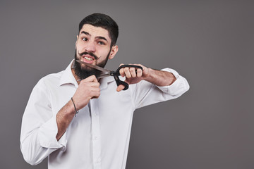 No more beard. Portrait of handsome young man cutting his beard with scissors and looking at camera while standing against grey background. New trend