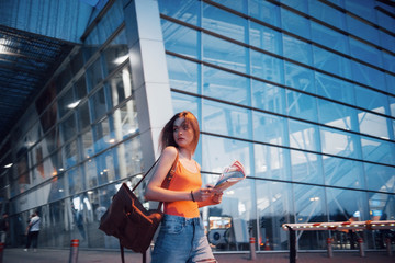 A young beautiful girl with a backpack behind her shoulders standing on the street near an airport. She just arrived from the rest and very happy