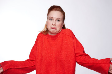 Fototapeta premium People, apathy and attitude concept. Questioned beautiful young woman shrugging with hesitation, feels doubt, has ginger hair, wears casual red oversized sweater, isolated against white background.