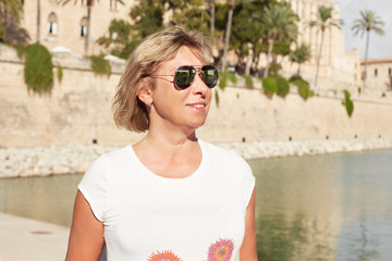 Good-looking Caucasian mature tourist female with blond hair wearing spectacles with sun protection and casual stylish t-shirt standing alone over city background, posing for photo, smiling gently.