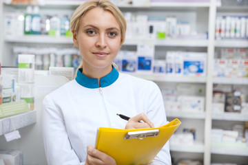 Confident female pharmacist taking notes on her clipboard, smiling to the camera joyfully. Lovely female chemist working at her drugstore, checking stock. Pharmacy, retail, medicament concept