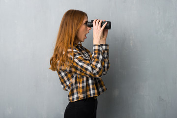 Young redhead girl over grunge wall and looking in the distance with binoculars