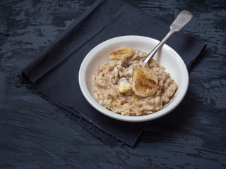 A deep ceramic plate with porridge and a spoon, a sprinkle of cinnamon and banana circles. Under the plate is a dark napkin.