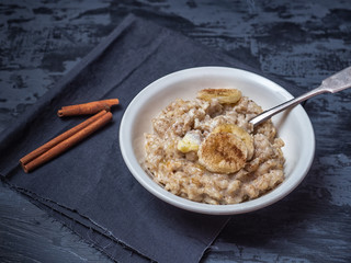 A plate of porridge with a sprinkling of cinnamon and banana circles. Under the plate is a dark napkin. Nearby are cinnamon sticks.