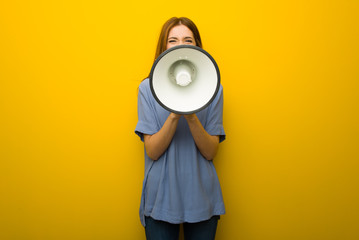 Young redhead girl over yellow wall background shouting through a megaphone to announce something