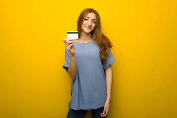 Young redhead girl over yellow wall background holding a credit card