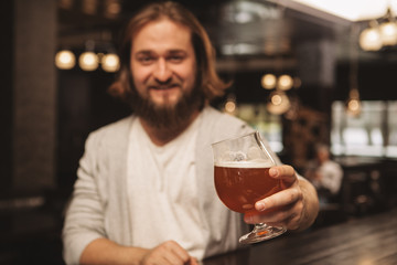 Selective focs on a glass of delicious craft beer in the hand of cheerful bearded man. Male customer enjoying weekend, having beer at local pub, copy space. Recreation, consumerism concept
