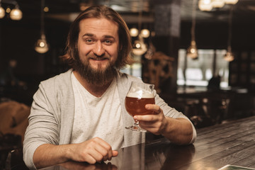 Cheerful young bearded man smiling happily to the camera, holding out glass of delicious craft beer, copy space. Happy man enjoying tasty beer at local pub. Excited male client resting at the bar