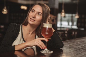 Lovely attractive woman relaxing at the pub, having delicious beer, looking away dreamily. Charming beautiful woman resting, drinking beer at the bar, thinking and smiling. Inspiration concept