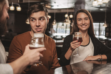 Happy beautiful young woman smiling to the camera, resting at the beer pub with her friends. Lovely female enjoying craft beer at the bar. Happiness, lifestyle, recreation concept