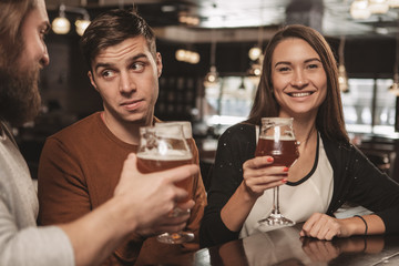 Happy beautiful young woman smiling to the camera, resting at the beer pub with her friends. Lovely female enjoying craft beer at the bar. Happiness, lifestyle, recreation concept