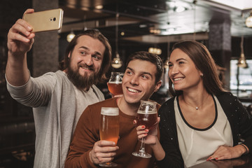 Group of friends having fun at beer pub together, taking selfies with smart phone. Bearded cheerful man using his phone, taking photos with his friends. Social media, technology concept
