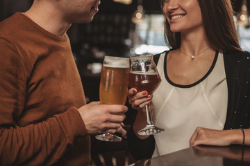 Selective focus on glasses of beer clinking in the hands of couple celebrating anniversary at restaurant. Cropped shot of a loving couple toasting with their beer glasses. Recreation, couples concept