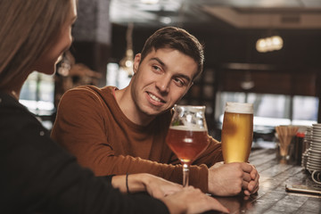 Handsome happy man talking to his girlfriend, while drinking craft delicious beer at the bar together. Attractive man flirting with his woman, having drinks together. Romance, dating, couple concept