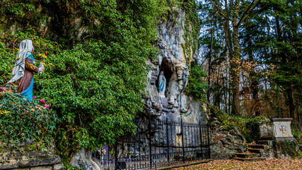 Statue of Bernadette praying to Our Lady of Lourdes in an outdoor chapel with a natural grotto,...