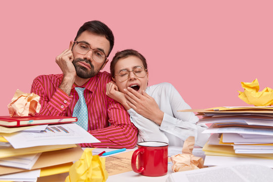 Tired woman and man coworkers have sleepy looks afer long hours working, surrounded with pile of paper documents, drink coffee, need to continue working on statistic report, isolated over pink wall