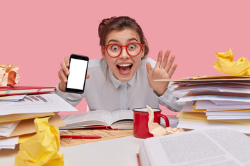 Smiling delighted European girl waves hand, holds mobile phone with mock up screen, being in high spirit, drinks hot beverage, surrounded with heap of documents, isolated over pink background