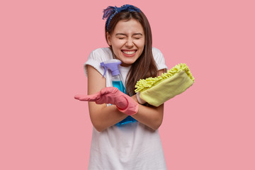 Positive female smirks face, has broad smile, white perfect teeth, carries rag with cleaning agent, being in good mood, giggles at something funny, isolated over pink background. Housework concept