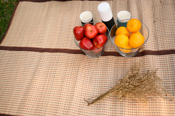 orange and apple in glass basket on mat picnic time . plastic glass on the mat