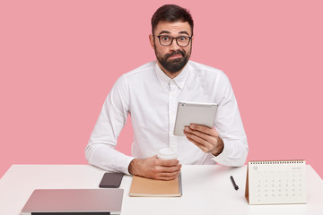 Uncertain male trader browses web page, checks mail in touchpad, frowns face, has thick stubble,...