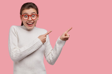 Studio shot of joyful woman points right with finger gun gesture, shows something wonderful against pink background, dressed in white casual outfit, isolated over pink studio wall. Check this