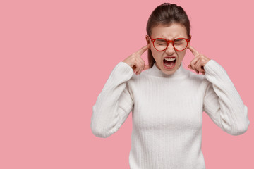 Terrible woman plugs ears with fore fingers, opens mouth widely, closes eyes, wears casual clothes,...