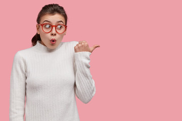 Isolated shot of surprised woman points with thumb, shows direction somewhere, dressed in casual...