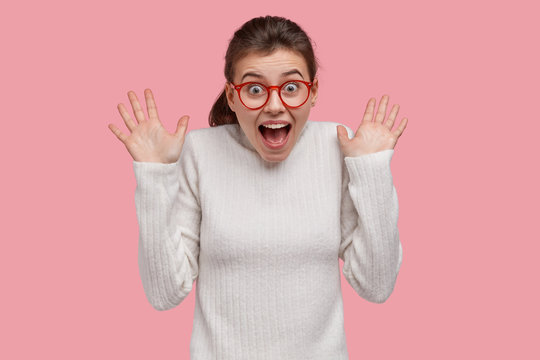 Photo of overemotive young woman reacts on sudden fake news, shows palms, keeps jaw dropped, wears transparent spectacles, dressed in white jumper, models over pink background. Emotions concept