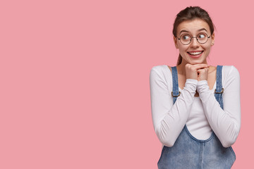 Optimistic glad woman with delighted look, keeps hands together under chin, concentrated aside,...