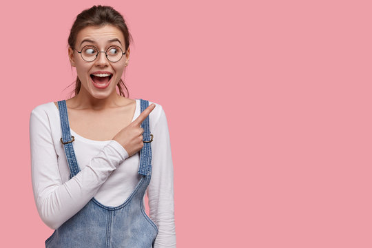 Photo of pleased brunette woman with joyful expression, indicates at upper right corner, wears optical glasses and denim outfit, shows free space for your promotional content. Wow, wonderful sales