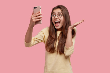 Happy woman spreads palm, smiles joyfully, holds modern cell phone in front, makes video call, glad to see old friend on distance, isolated over pink background. People, technology, emotions concept