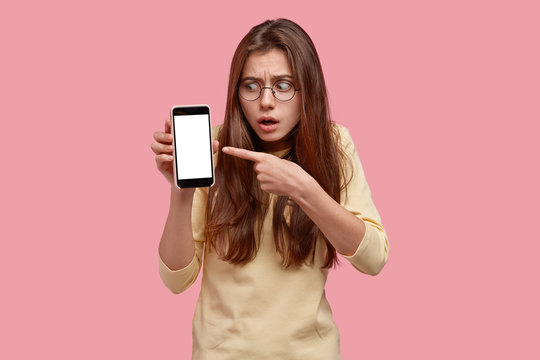 Worried displeased woman has long hair, wears round glasses, points with index finger at smart phone with empty screen, demonstrates something strange, isolated over pink background. Technology