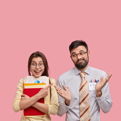 Vertical shot of happy girl points at hesitant guy who spreads hands, laughs at nerd, carry papers, isolated over pink background with copy space upwards for your advertisement or promotion.