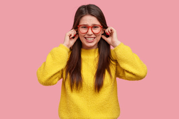 Studio shot of annoyed woman plugs ears with fingers, expresses negativeness, clenches teeth, wears casual yellow clothes, poses against pink background, stops annoying sound. Negative emotions
