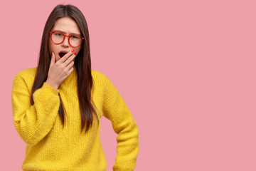 Photo of displeased brunette tired woman yawns and covers mouth with palm, wears optical glasses and yellow sweater, stands against pink background with blank space for your promotional content